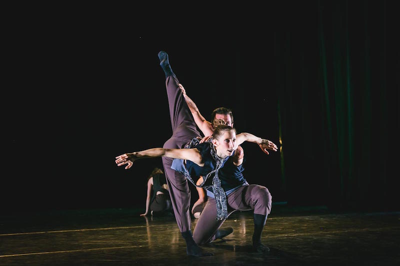 A female dancer executes a penchee while a male dancer holds onto her leg